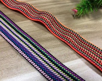 Può includere: Due bande elastiche intrecciate colorate con un motivo geometrico. La banda in alto è arancione, nera, verde e bianca. La banda in basso è viola, rosa, verde, nera e bianca.