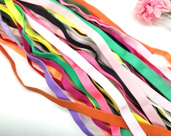 May include: A collection of colorful elastic ribbons in various shades, including pink, green, orange, and purple. The ribbons are arranged diagonally, creating a vibrant display. A few ribbons are black and white. A pink flower is in the background.