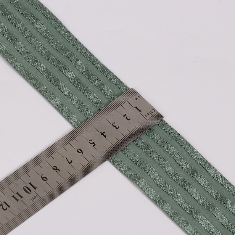 Peut inclure: Un ruban &eacute;lastique vert sauge pr&eacute;sentant une s&eacute;rie de rayures parall&egrave;les, en relief et scintillantes sur toute sa longueur. Une r&egrave;gle en acier inoxydable est plac&eacute;e en diagonale sur le ruban, indiquant une largeur d'environ 3,8 cm.