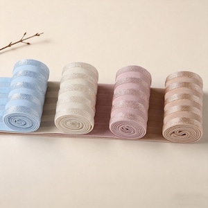 Peut inclure: Quatre rouleaux de bandes &eacute;lastiques en tissu c&ocirc;tel&eacute; dans les tons bleu clair, cr&egrave;me, vieux rose et beige. Chaque rouleau pr&eacute;sente un motif r&eacute;p&eacute;titif de rayures horizontales alternant entre mat et m&eacute;tallique brillant sur la surface textur&eacute;e.