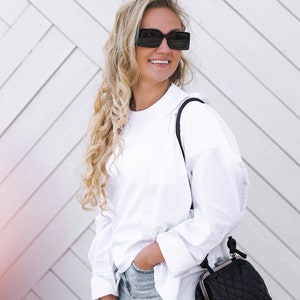 A stylish woman wearing sunglasses, a white blouse, and jeans poses with a black quilted handbag outdoors.