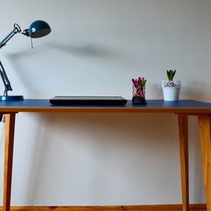 May include: A blue desk with a wooden frame and a blue lamp. A laptop, a cup of pens, and a potted plant are on the desk.