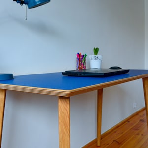 May include: A blue desk with a light wood frame and legs. The desk has a laptop, a potted plant, and a desk lamp on it.