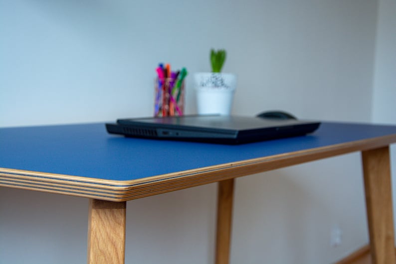 May include: A blue desk with a wooden frame and a laptop on top. The desk has a light wood edge and is sitting on a white floor.