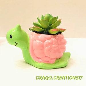 Puede incluir: Un macetero de tortuga de cerámica verde y rosa con una planta suculenta en su interior. La tortuga tiene una cara sonriente y un solo ojo. El macetero es perfecto para plantas pequeñas y suculentas.