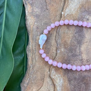 Può includere: Una collana di perline rosa con un ciondolo bianco irregolare unico. La collana è composta da piccole perline rotonde rosa chiaro. Il ciondolo è di colore bianco perla chiaro ed è attaccato con una piccola chiusura dorata.