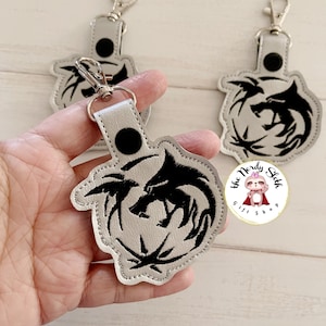 May include: A silver leather keychain with a black embroidered wolf design. The keychain has a silver metal ring and a black button on the back. The text "the Nerdy Sloth Gift Shop" is visible in the image.