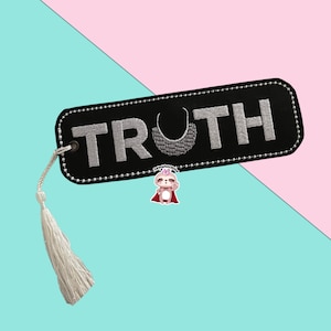 May include: Black bookmark with white embroidered text "TRUTH" and a small, embroidered sloth wearing a red cape. The bookmark has a white tassel.