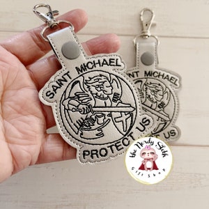 May include: A white leather keychain with a black embroidered image of Saint Michael the Archangel. The keychain features the text "Saint Michael Protect Us".