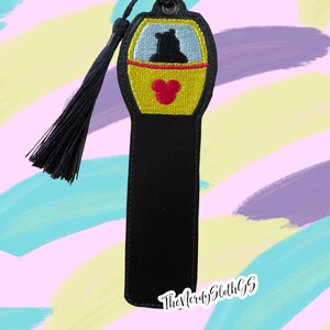 May include: Black leather bookmark with a yellow and blue embroidered design of a ride at a theme park. The design features a black silhouette of a person in a yellow car with a red Mickey Mouse head.