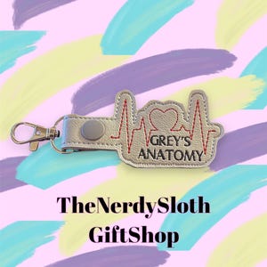 May include: A silver key chain with a red heart and a red heartbeat line. The keychain has the text "GREY'S ANATOMY" on it.