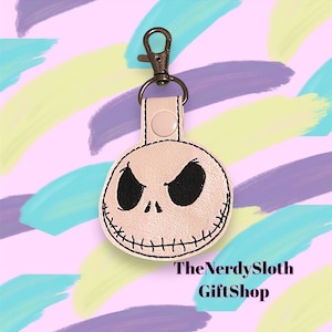 May include: A pink leather keychain with a black and white embroidered Jack Skellington face. The keychain has a silver metal key ring and a silver metal clasp. The text "The Nerdy Sloth GiftShop" is printed below the keychain.