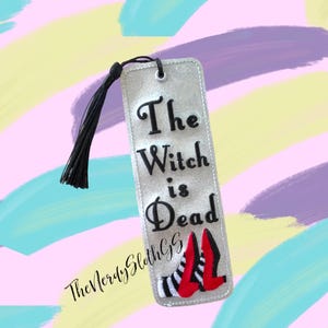 May include: A silver bookmark with a black tassel. The bookmark has black embroidery that reads "The Witch is Dead" with a pair of red and black shoes below the text.