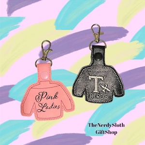 May include: Two keychains shaped like sweaters. One is pink with the text "Pink Ladies" embroidered on it. The other is black with the text "T-Birds" embroidered on it.