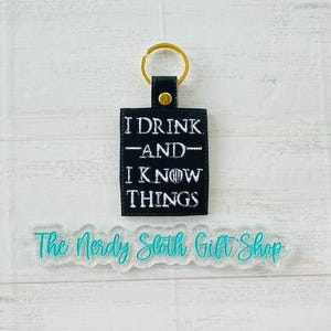 May include: Black leather keychain with a white embroidered saying "I drink and I know things". The keychain has a gold metal ring. The background is a light wood surface. The text "The Nerdy Sloth Gift Shop" is printed on a clear, glittery surface.