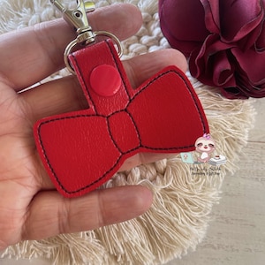 May include: Red leather bow keychain with a silver metal clasp. The bow has a red button in the center.