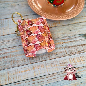 May include: A white hand sanitizer holder with a colorful pattern of pastries and sweets. The holder has a gold key chain attached to it. The holder is on a wooden surface.