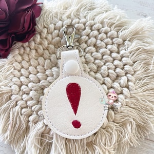 May include: White leather keychain with a red exclamation point embroidered on a round tag. The keychain has a silver metal ring.