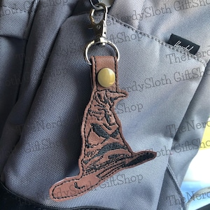 May include: A brown leather keychain with a black embroidered image of the Sorting Hat from the Harry Potter series. The hat is sitting on a gray backpack.