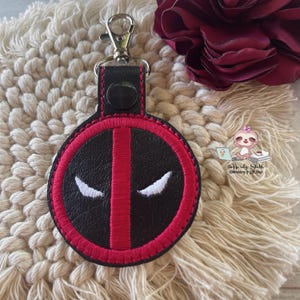 May include: A black and red round keychain with a white embroidered design of Deadpool's eyes. The keychain is attached to a silver metal keyring.