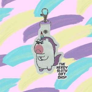 May include: White unicorn keychain with a grumpy expression, rainbow horn, and purple mane. The keychain has a silver key ring and the text "The Nerdy Sloth Gift Shop" is embroidered below the unicorn.