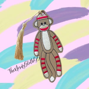 May include: A brown sock monkey bookmark with red and white stripes, wearing a red and white hat. The bookmark has a gold tassel.