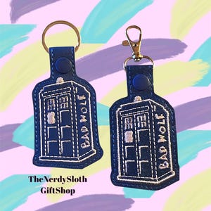 May include: Two navy blue keychains shaped like a police call box, with the words "BAD WOLF" embroidered in white. One keychain has a gold ring, the other a gold clasp. The text "The Nerdy Sloth GiftShop" is at the bottom.