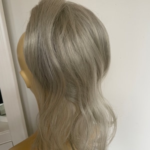 Könnte beinhalten: Eine silbergraue Clip-in-Ponytail-Haarverlängerung. Das Haar ist wellig und zu einem Pferdeschwanz gestylt.