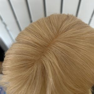 Puede incluir: Primer plano de una peluca rubia con una raya visible. El cabello es de un rubio claro y cálido y parece estar peinado de forma natural. La construcción de la peluca es visible en la raya.