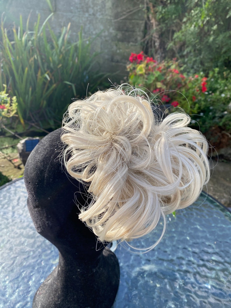 May include: A blonde hairpiece in a messy bun style. The hairpiece is on a black mannequin head.