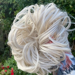 May include: A white blonde hair bun extension. The hair is curled and styled in a messy bun.