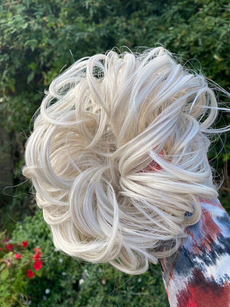 May include: A white blonde hair bun extension. The hair is curled and styled in a messy bun.
