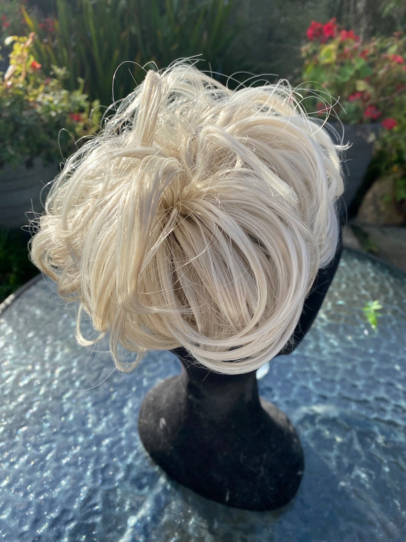May include: A blonde hair bun extension on a black mannequin head. The hair is styled in a messy bun.