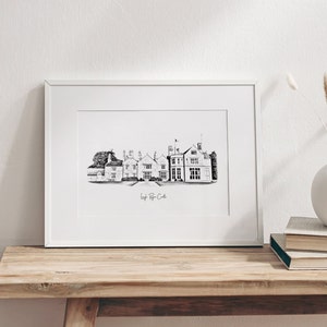 May include: A framed black and white architectural print of a building, likely a castle or manor house, with the text "Luton Hoo Castle" below the image. The print is in a white frame and rests on a wooden table.