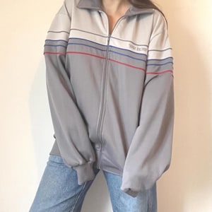 May include: A grey zip-up jacket with a white, blue and red striped design. The jacket has the text "VOGUE SELECT" on the right side of the chest.
