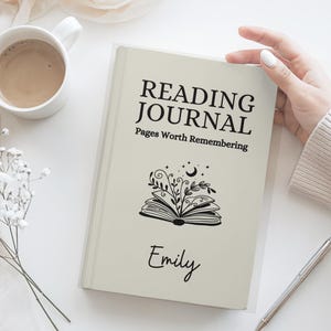 May include: A cream-colored reading journal with the text "READING JOURNAL" and "Pages Worth Remembering." A floral and moon illustration is below the text, with the name "Emily" below the illustration. A pen and a cup of coffee are also in the image.