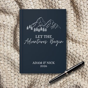 Könnte beinhalten: Ein marineblaues Notizbuch mit weißen Berg- und Baumillustrationen und dem Text "LET THE Adventures Begin". Das Notizbuch enthält auch die Namen "ADAM & NICK" und das Jahr "2026". Ein schwarzer Stift liegt auf der strukturierten Oberfläche.