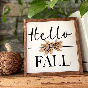 May include: A wooden sign with the text "Hello Fall" in black lettering. The sign has a white background and is decorated with two brown wooden leaves.