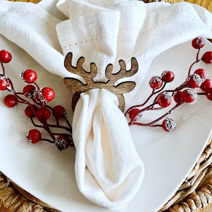 May include: A white linen napkin is held by a wooden reindeer napkin ring on a white plate. Red berries with silver glitter accents are arranged around the plate, which sits on a woven placemat. The setting suggests a festive holiday theme.