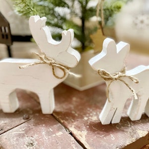 May include: Two white wooden reindeer figurines with a rustic aesthetic. Each reindeer has a simple design and is adorned with a twine bow around its neck. The figurines are placed on a weathered wooden surface, suggesting a holiday or seasonal theme.