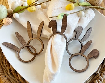 Puede incluir: Cuatro anillos de servilleta de madera en forma de conejo en tono madera marrón. Los anillos están colocados en un plato blanco con una servilleta blanca. La servilleta está decorada con una rama de hojas verdes y huevos de Pascua amarillos y blancos.