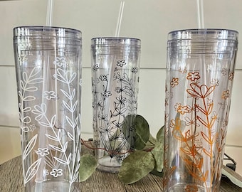 May include: Three clear plastic tumblers with lids and straws, each decorated with floral designs. One has white flowers, another black, and the third has orange flowers. The tumblers are cylindrical and sit on a wooden surface.