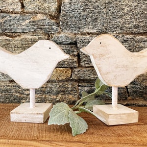 May include: Two white wooden bird figurines with a distressed finish. Each bird is perched on a small, square base and has a simple, minimalist design. The birds are approximately 6 inches tall, and are suitable for home decor.