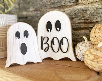 May include: Two wooden ghost decorations with black eyes and mouths. One ghost has an open mouth, and the other says "BOO" in black script. The ghosts are white with a distressed finish, and are part of a Halloween theme.