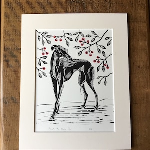 May include: Black and white linocut print of a greyhound dog beneath a cherry tree. The dog is in profile, with detailed linework. The cherry tree branches have red berries. The print is matted in a cream-colored mat.