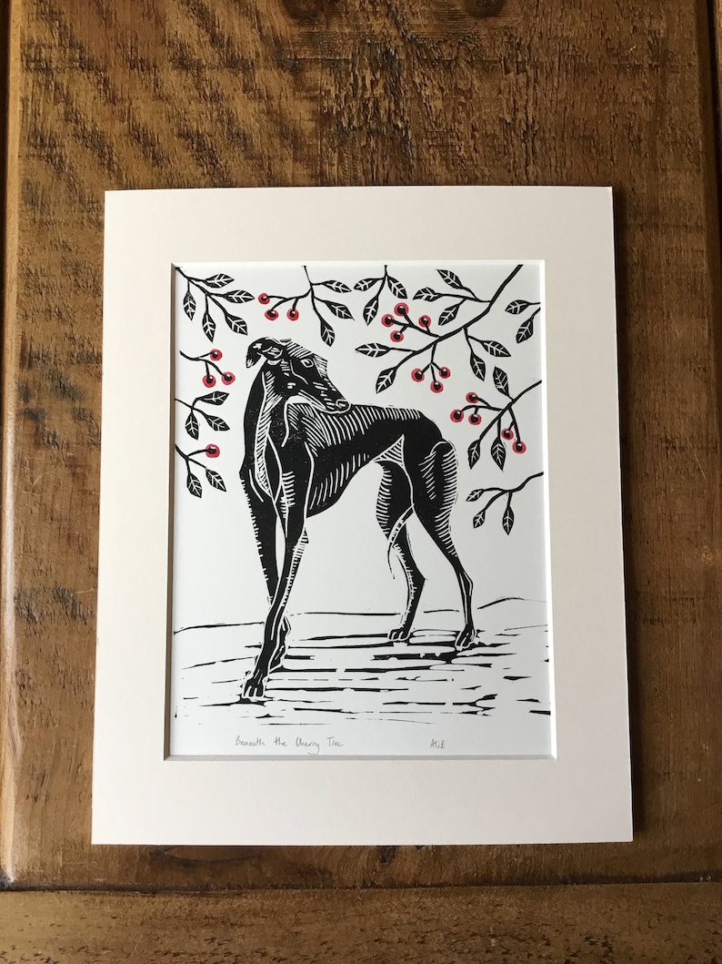 May include: Black and white linocut print of a greyhound dog beneath a cherry tree. The dog is in profile, with detailed linework. The cherry tree branches have red berries. The print is matted in a cream-colored mat.