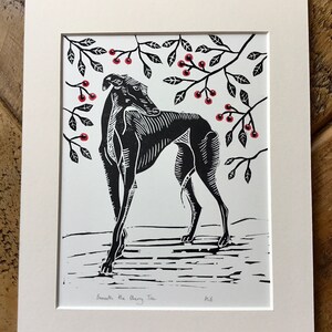 May include: Black and white linocut print of a dog beneath a cherry tree. The dog is stylized with detailed linework. Red cherries and black leaves are in the branches above. The print is matted in a white frame. The text "Beneath the Cherry Tree" is below the image.