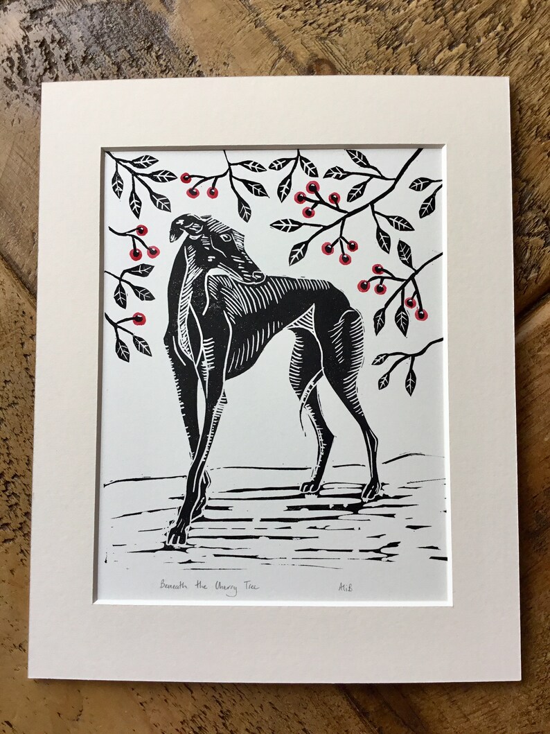 May include: Black and white linocut print of a dog beneath a cherry tree. The dog is stylized with detailed linework. Red cherries and black leaves are in the branches above. The print is matted in a white frame. The text "Beneath the Cherry Tree" is below the image.
