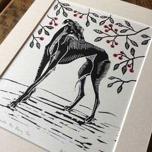 May include: Black and white linocut print of a dog beneath a cherry tree. The dog is arched, with branches of leaves and red cherries above. The print is on white paper, matted, and framed.