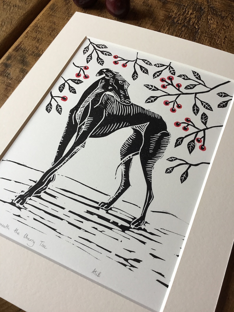 May include: Black and white linocut print of a dog beneath a cherry tree. The dog is arched, with branches of leaves and red cherries above. The print is on white paper, matted, and framed.